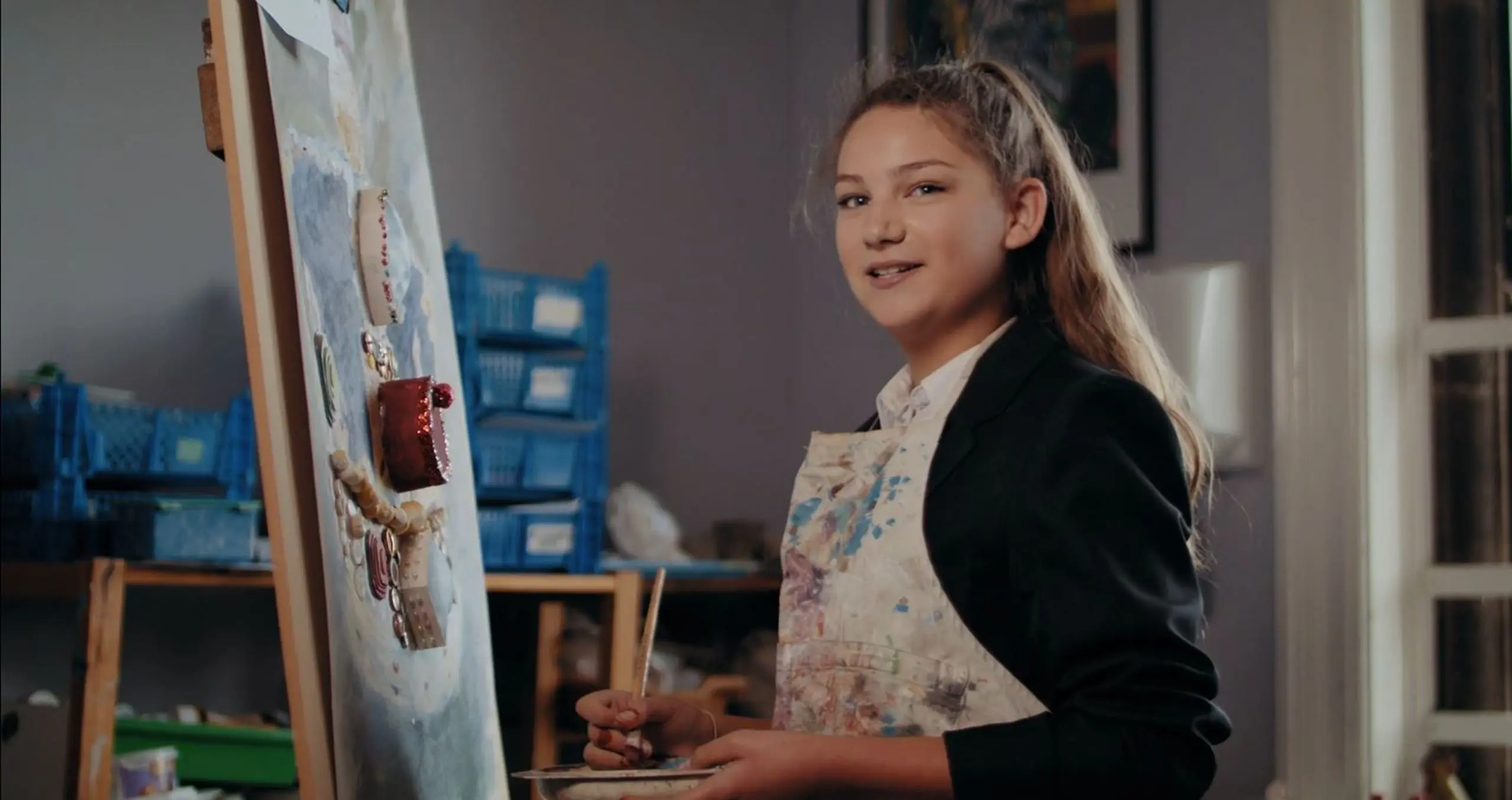 A female Moon Hall School student wearing an art apron, smiling while working on a canvas painting in an art classroom.