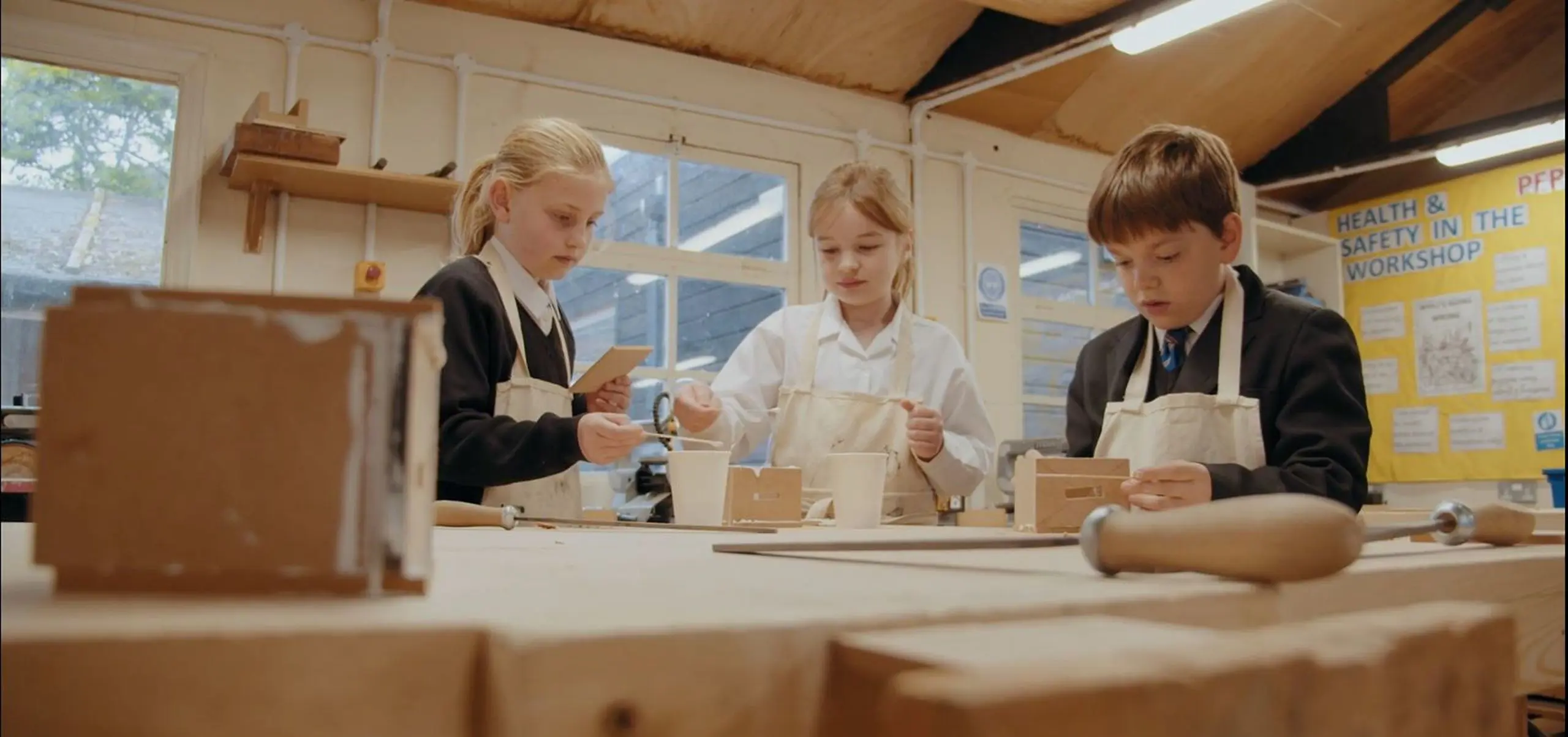 Three pupils wearing aprons working on woodwork projects in a school design and technology workshop