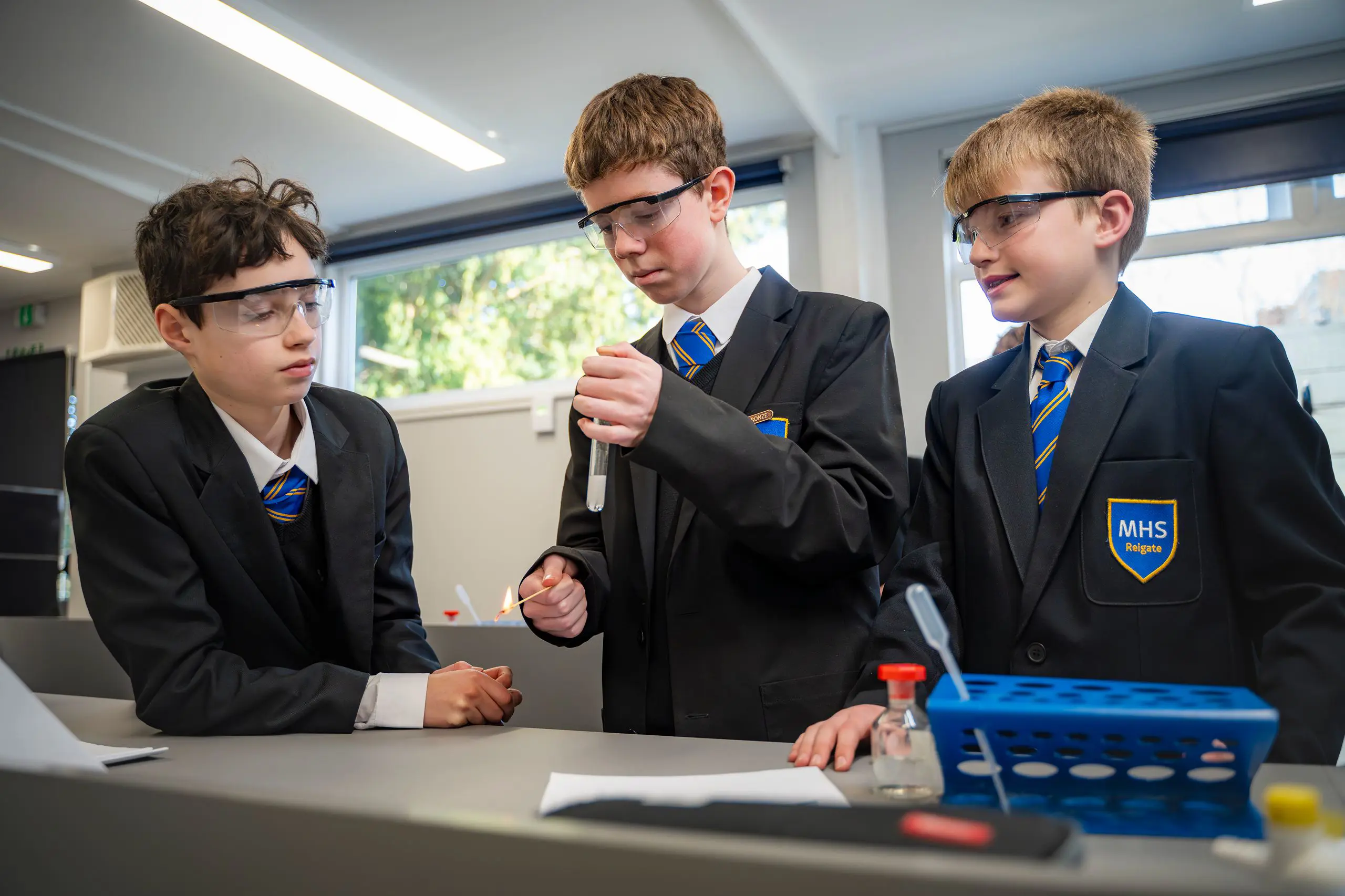 Three male pupils in school uniform wearing protective safety goggles during a science experiment in a classroom