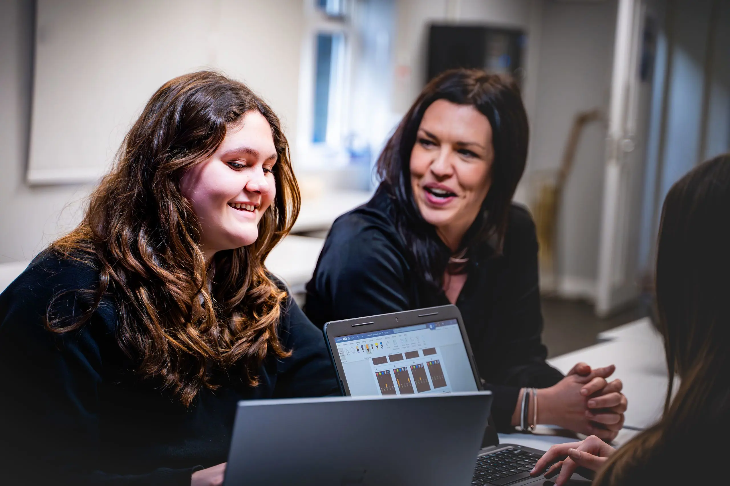Teacher smiling and talking with a female pupil working on a laptop