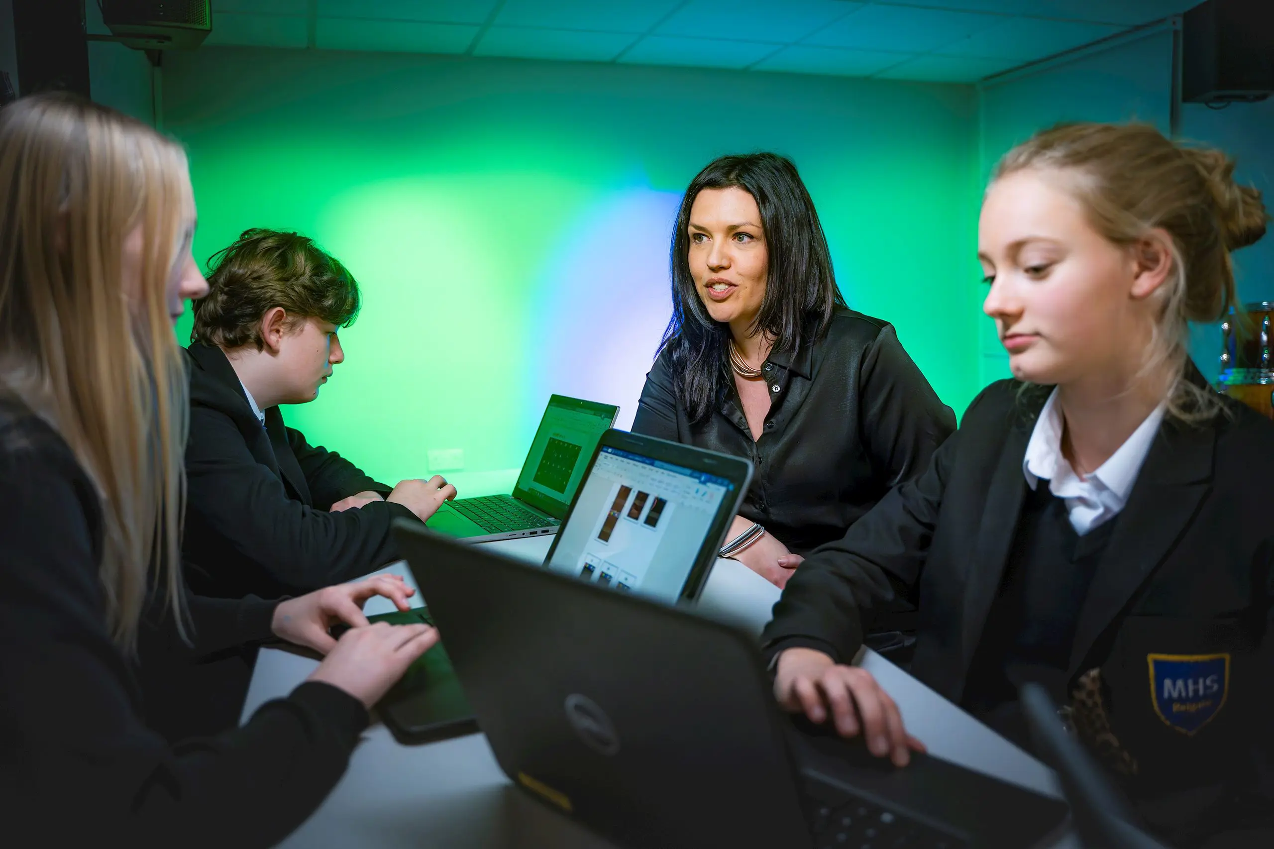 Teacher seated at a computer table assisting three pupils working on laptops