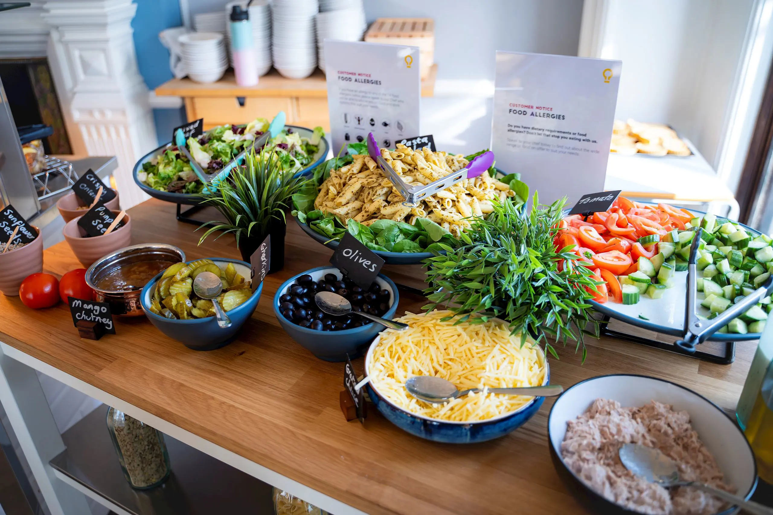 Selection of fresh salad bar items, bowls of grated cheese, olives, sweetcorn, pasta, and chopped vegetables displayed on a wooden table