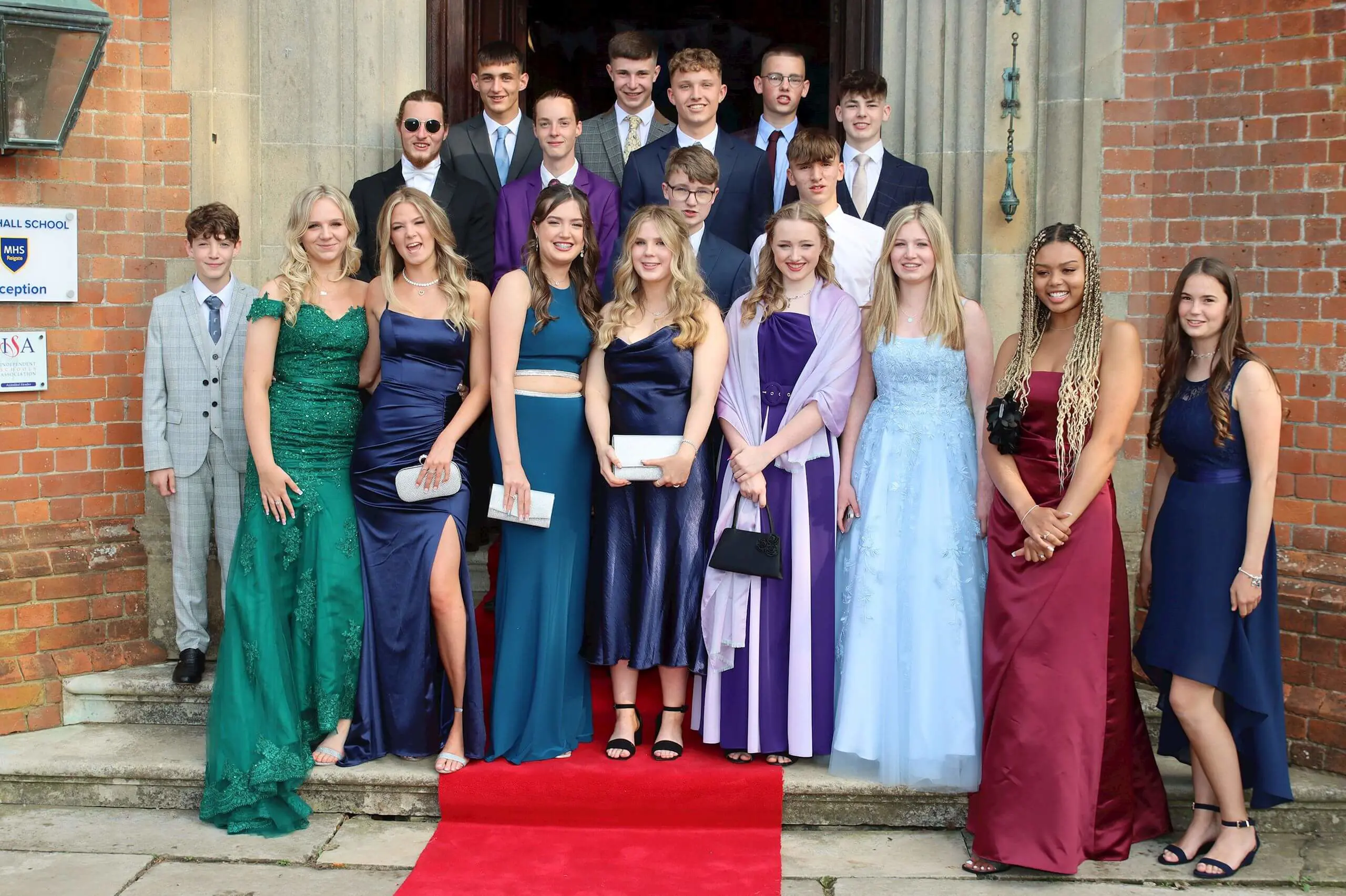 Group of Moon Hall School pupils dressed in formal prom attire posing together on brick steps with a red carpet