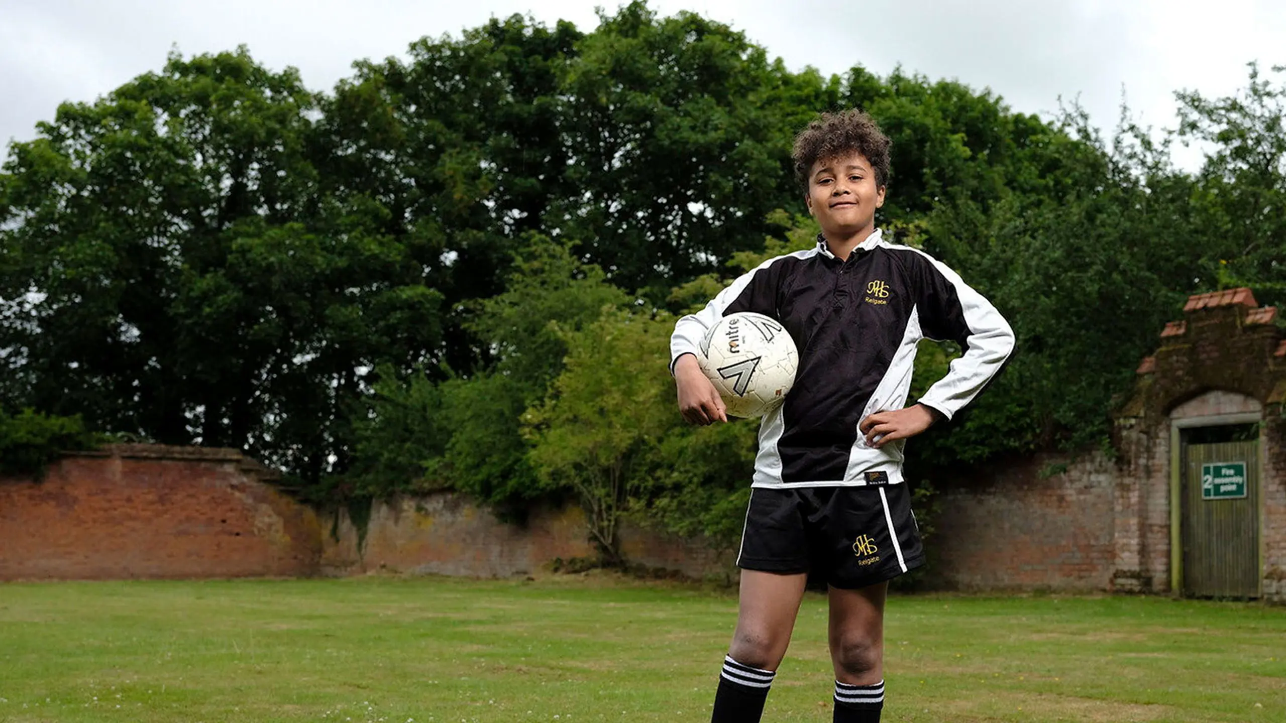 Young male student in sports kit holding a rugby ball on the school playing fields