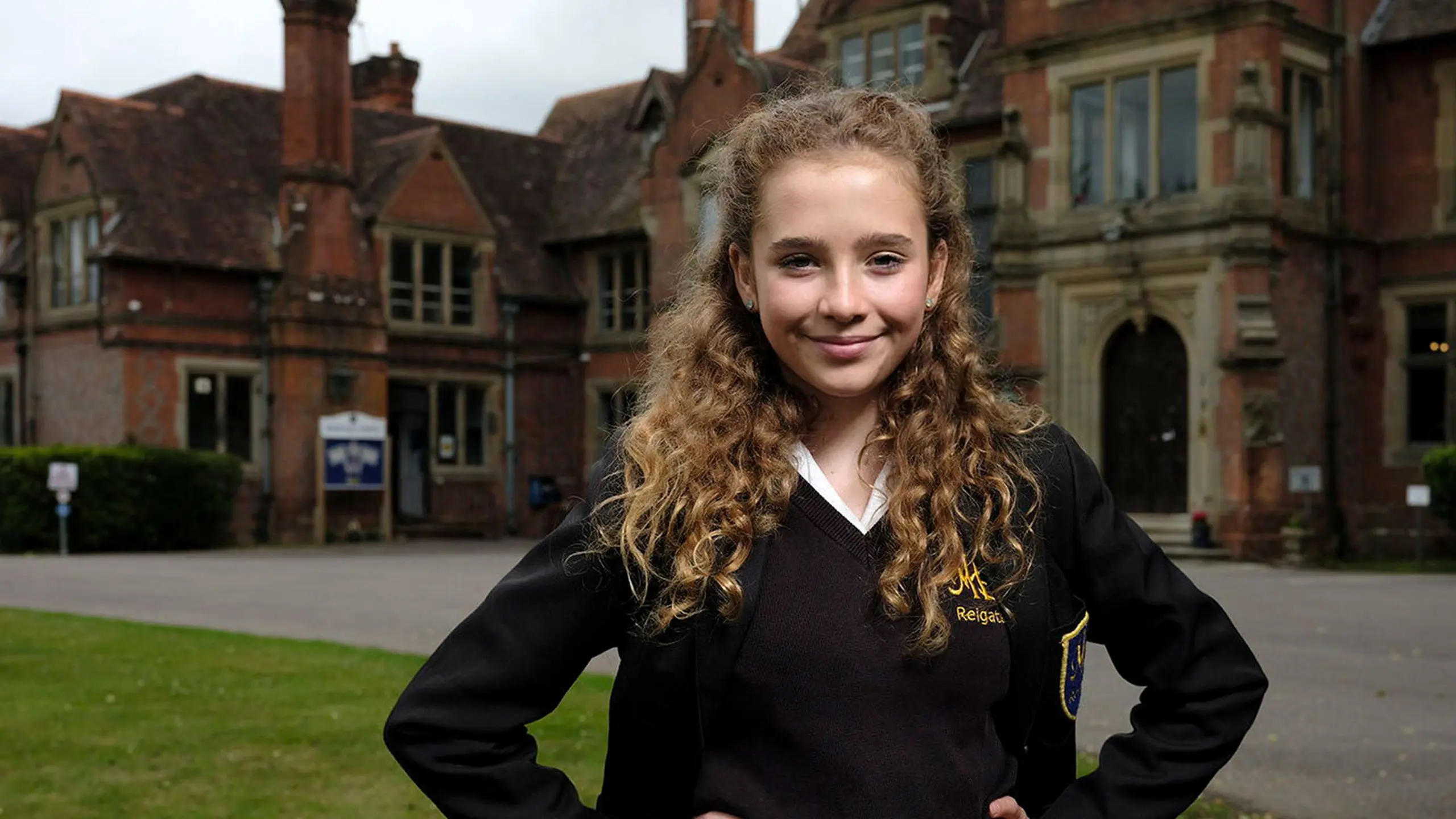 Smiling female pupil in school uniform standing in front of the historic Moon Hall School building