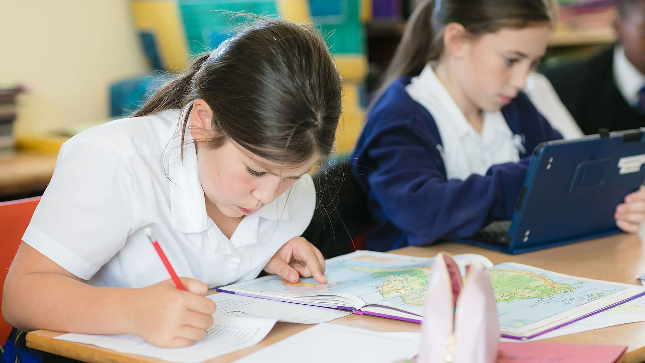 Two young female students sitting at a desk in a classroom, with one writing in an exercise book and the other using a tablet computer