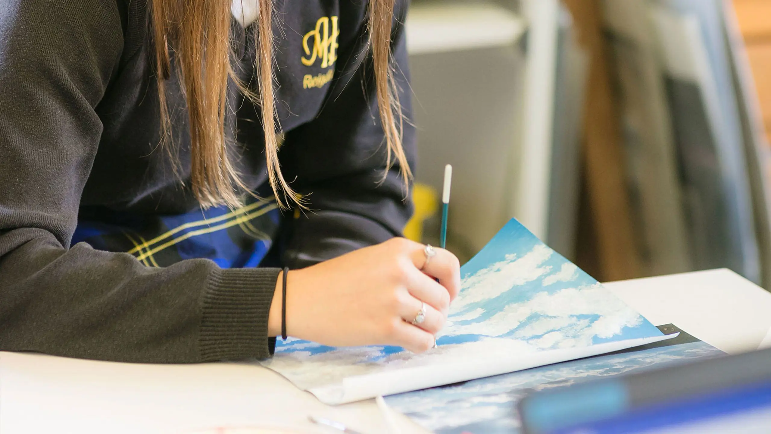 Close-up of a student in a Moon Hall School jumper holding a paintbrush and creating artwork at a classroom desk