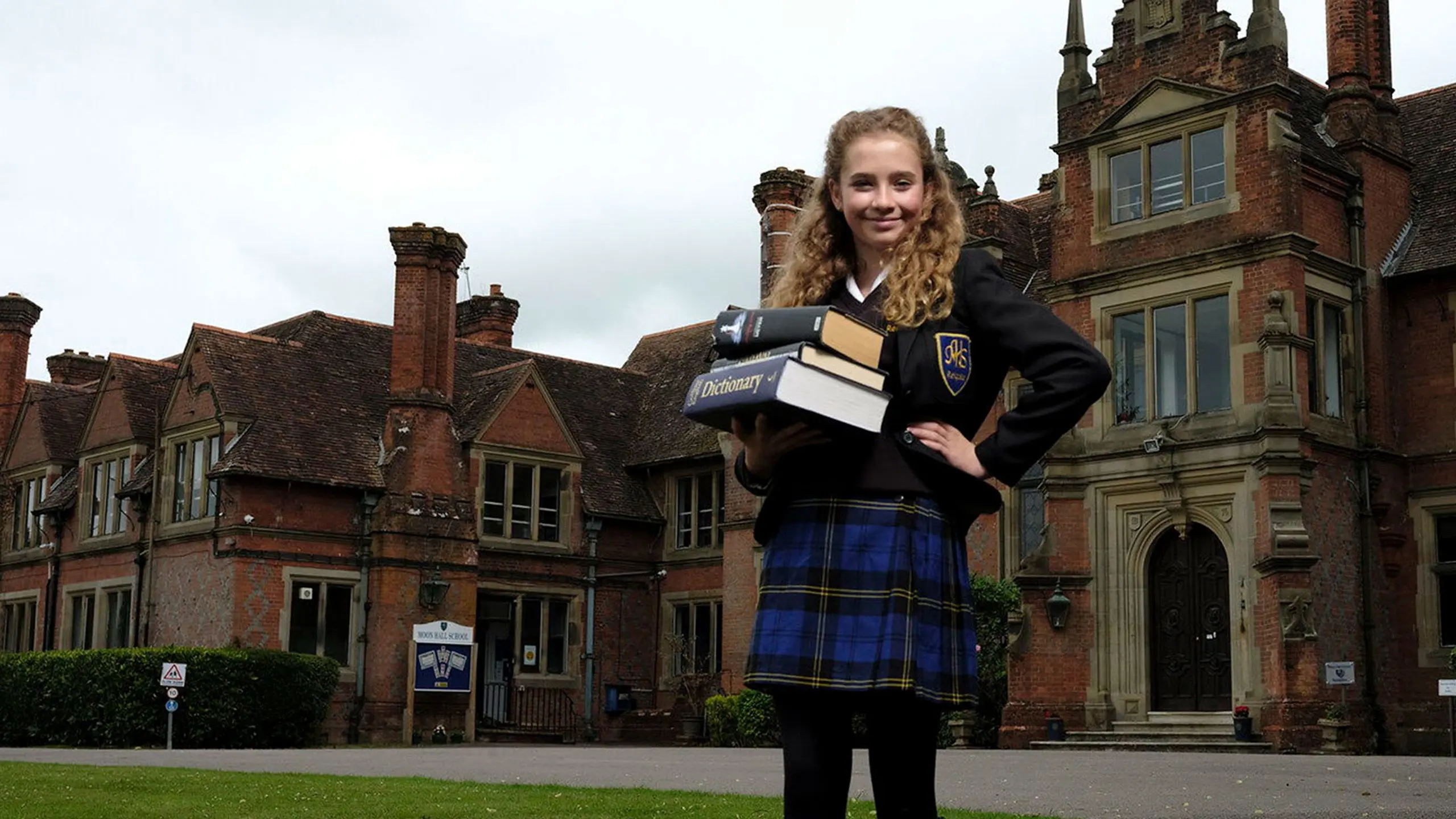 Female pupil holding a stack of textbooks outside the historic Moon Hall School building