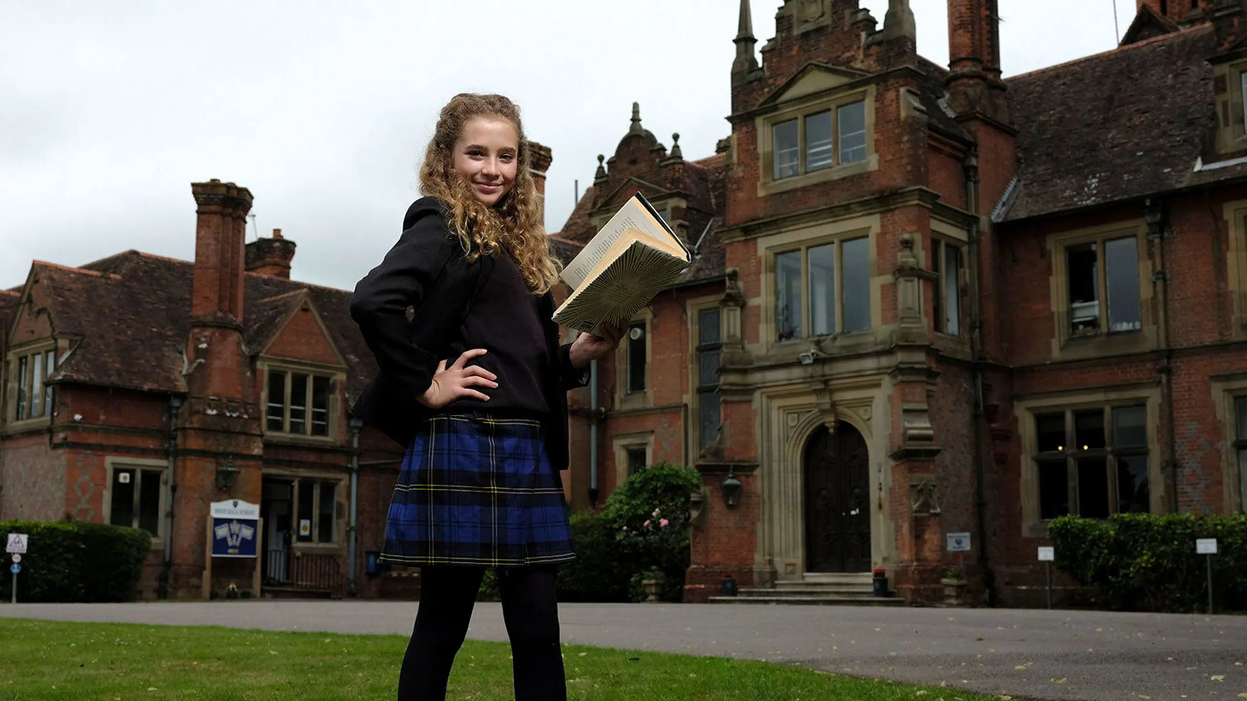 Female pupil holding a book and posing on the school lawn in front of the main Moon Hall School building