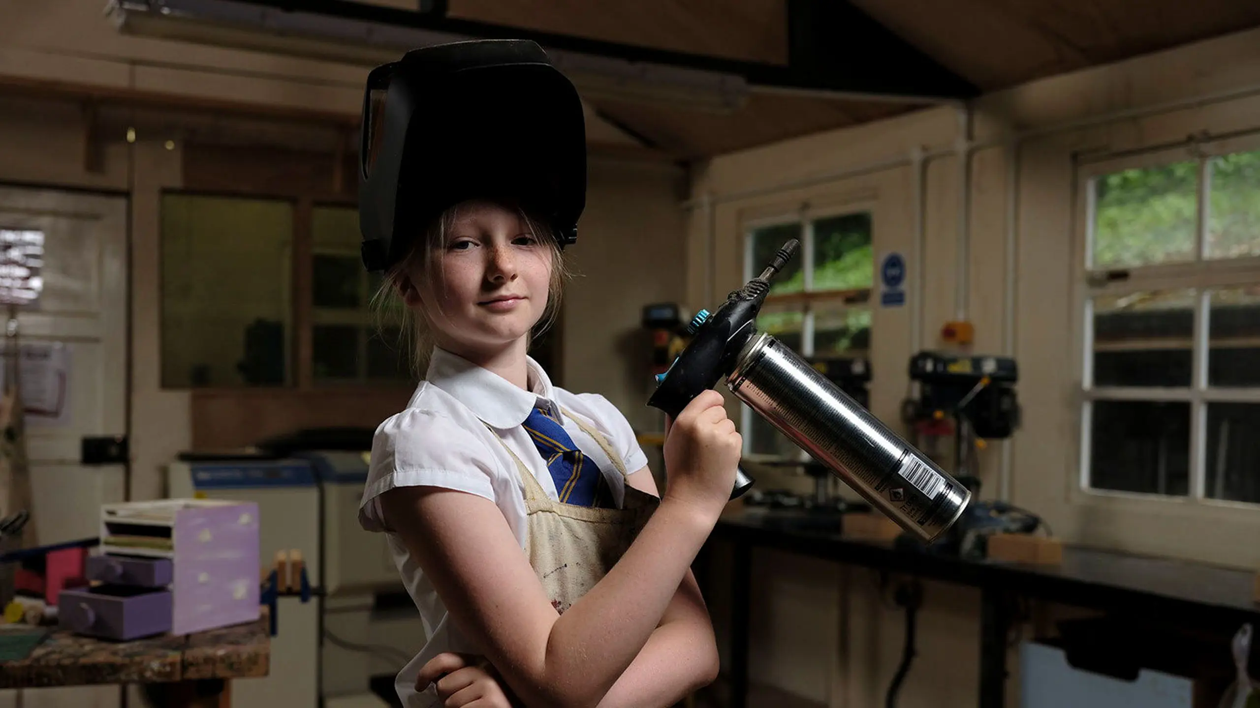 Young female student wearing a school uniform, apron, and protective welding visor, holding a piece of workshop equipment in a design technology room